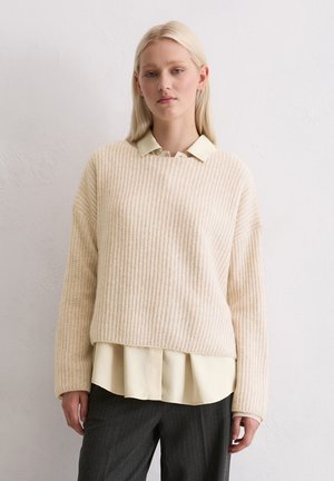 Maglione beige a coste con scollo rotondo indossato sopra una camicia color giallo chiaro con colletto. L'orlo inferiore è leggermente accorciato e le maniche sono ampie.