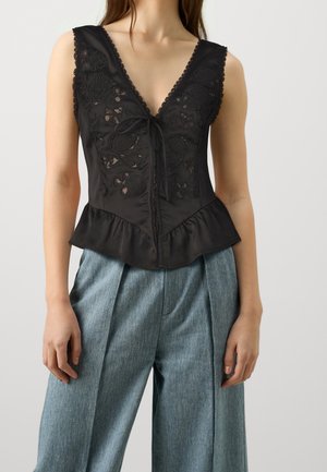 Femme portant un top sans manches noir à lacets brodé avec un ourlet volanté et un pantalon en denim léger bleu clair, le tout sur fond uni.