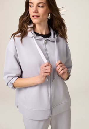Felpa con zip di colore grigio chiaro, dotata di cappuccio, vestibilità rilassata, maniche corte e lacci. Tessuto morbido con una leggera texture. Dettagli della zip in argento.