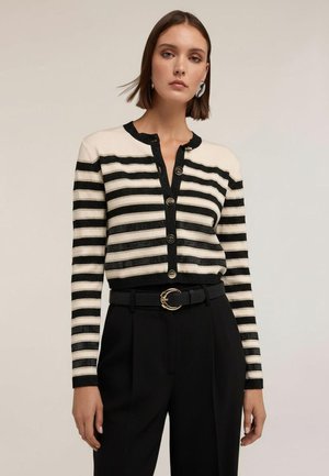 Femme portant un cardigan boutonné rayé noir et blanc, un pantalon noir taille haute avec une ceinture noire ornée d'une boucle dorée.