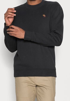 Mann trägt schwarzen Sweatshirt mit langen Ärmeln und kleinem gesticktem Elch-Logo auf der linken Brust, kombiniert mit beigen Hosen, und richtet seinen Ärmel.
