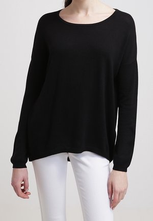 Kvinde iført en løs sort langærmet sweater og hvide bukser, stående mod en ensfarvet lys baggrund.