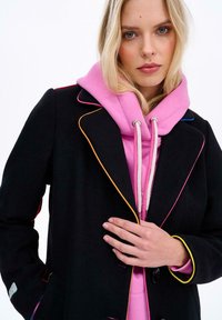 Žena s blond vlasmi, oblečená v ružovej mikine pod čiernym kabátom s farebným lemovaním, držiaca klopu kabátu jednou rukou.