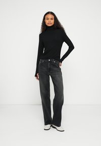 Bluză neagră cu mâneci lungi și guler înalt, combinată cu blugi largi, de culoare gri închis. Modelul poartă cizme de gleznă de culoare deschisă. Design simplu, modern.