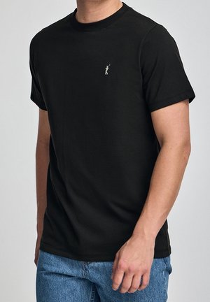 Man draagt een simpel zwart T-shirt met korte mouwen en een klein golferlogo op de borst, gecombineerd met blauwe spijkerbroek.