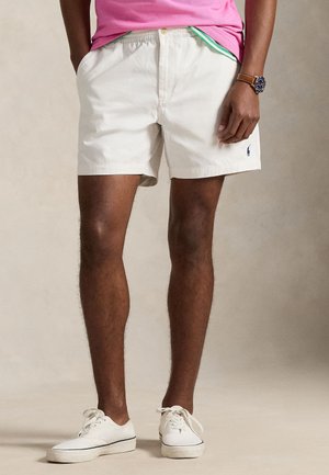 Homme portant un short blanc, des baskets blanches et une chemise rose, debout les mains dans les poches devant un fond uni.