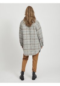Gråternet oversized skjorte med brune accenter, lange ærmer, buet kant, parret med brune bukser og sorte ankelstøvler.