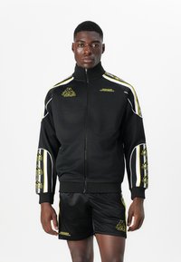 Black athletic zip-up jacket with yellow accents and logo, worn with shiny black shorts. Features bold side stripes and fitted design.