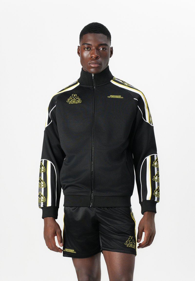 Black athletic zip-up jacket with yellow accents and logo, worn with shiny black shorts. Features bold side stripes and fitted design.
