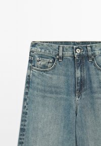 Svetle modré džíny z denimu s předním zapínáním na knoflíky, poutky na opasek a předními kapsami, včetně malé kapsičky na mince, na bílém pozadí.