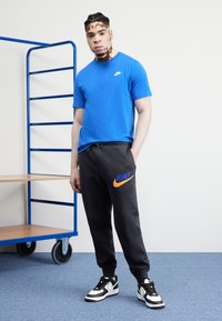 T-shirt blu a girocollo, pantaloni sudati neri con logo Nike arancione e blu, e sneakers in bianco e nero. Tessuto liscio; look casual e atletico.
