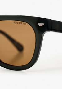 Gafas de sol negras con lentes de tono marrón, con forma rectangular, texto del logotipo en la montura y un sutil acento metálico en la bisagra.