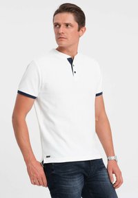 Bílá polokošile s krátkým rukávem, s knoflíkovou légou, tmavě modrým límečkem a manžetami, textura látky, a s logem na lemu.