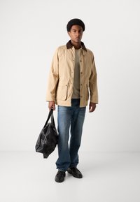 Beige jakke med brun krage, knaplukning og to frontlommer, parret med en grå skjorte, blå jeans og sorte sko; holder en sort taske.