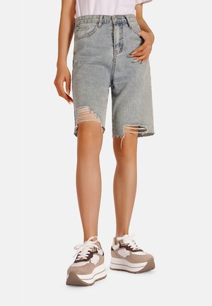 Shorts in denim blu distressed con orli sfilacciati, vita alta e dettagli strappati visibili, abbinati a sneakers chunky beige e bianche.