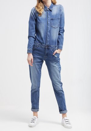 Femme portant une combinaison en jean bleu avec les poignets retroussés et des baskets blanches, debout avec une main dans la poche devant un fond uni.