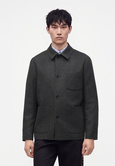 Jeune homme portant une veste à carreaux gris foncé avec boutons sur une chemise bleu clair à col, debout devant un fond blanc uni.