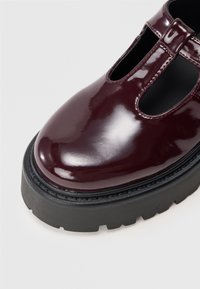 Zapato Mary Jane de charol burdeos con gruesa suela de plataforma negra y diseño de correa en T sobre un fondo blanco.