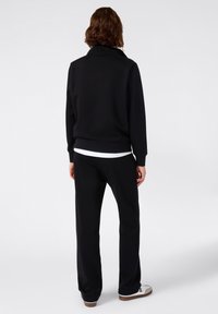 Felpa nera con collo alto e polsini a coste, abbinata a pantaloni neri coordinati. Camicia bianca visibile sotto. Calzature casual.
