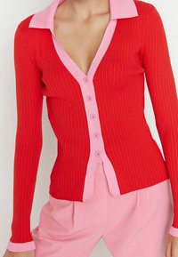 Frau trägt eine gerippte rote Strickjacke mit pinkfarbenen Kanten und Knöpfen über pinkfarbenen, hoch taillierten Hosen, steht vor einem einfarbigen Hintergrund.