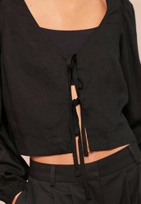 Afkortet sort bluse med V-halsudskæring, bindsnøring og puffede ærmer. Stoffet ser tekstureret ud med et let design.