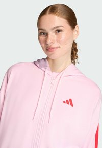 Ružová mikina na zips vyrobená z mäkkej látky s textúrovaným povrchom. Má červený akcent a logo na hrudi. Ľahký dizajn s kapucňou.