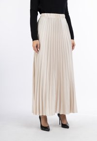 Plisseret midi-nederdel i beige, lavet af let stof. Har en elastisk talje og er stylet med sorte højhælede sko.