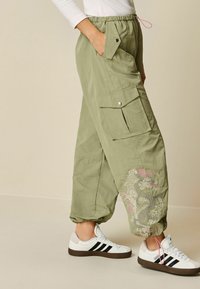 Pantalons cargo en tissu vert clair avec une taille élastique, des poches et une broderie florale rose sur le bas de la jambe, assortis à des baskets blanches.