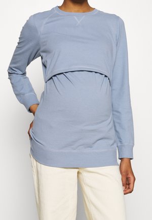 Sweatshirt de maternité à manches longues bleu clair avec un superposé court, des coutures texturées et des poignets côtelés, associé à un pantalon blanc cassé.