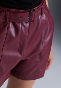 Burgundy leather shorts with a smooth texture, featuring a belted high waist and side pockets, styled with subtle pleats and silver hardware.