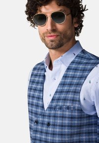 Niebieska kamizelka w kratę z podwójnym zapięciem, w zestawie z jasnoniebieską koszulą z drobnym wzorem łodzi żaglowych. Okrągłe okulary przeciwsłoneczne dopełniają całość.