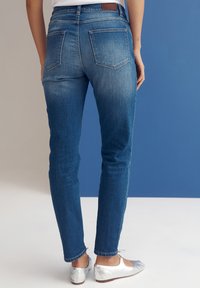 Skinny blue jeans with a faded wash, five-pocket design, and a brown leather patch at the waist, shown from the back.