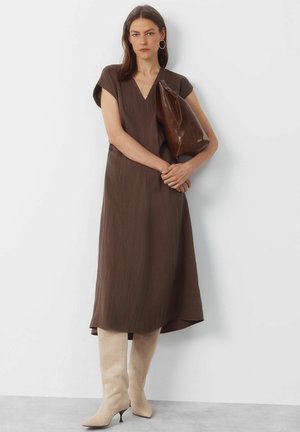 Femme portant une robe midi marron, des bottes beige montantes jusqu'aux genoux, des boucles d'oreilles créoles en or, tenant une grande pochette en cuir marron contre un mur blanc.