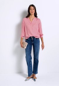 Blusa rosa chiaro a maniche lunghe con scollatura a V e dettagli di cucitura, abbinata a jeans blu a gamba dritta e ballerine argentate a punta. Tiene una pochette intrecciata.