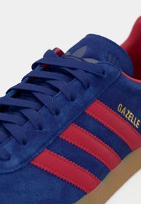 Close-up de um ténis de camurça azul com riscas vermelhas, interior vermelho, atacadores azul-marinho e o texto "GAZELLE" em dourado na lateral.