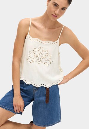 Mujer con una camiseta blanca sin mangas con bordado floral calado y bordes festoneados, combinada con pantalones cortos vaqueros azules y un cinturón trenzado marrón.