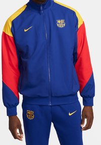 Chándal Nike azul y rojo con acentos amarillos. La chaqueta tiene un cierre de cremallera en la parte delantera, puños acanalados y el logo del FC Barcelona en el pecho.