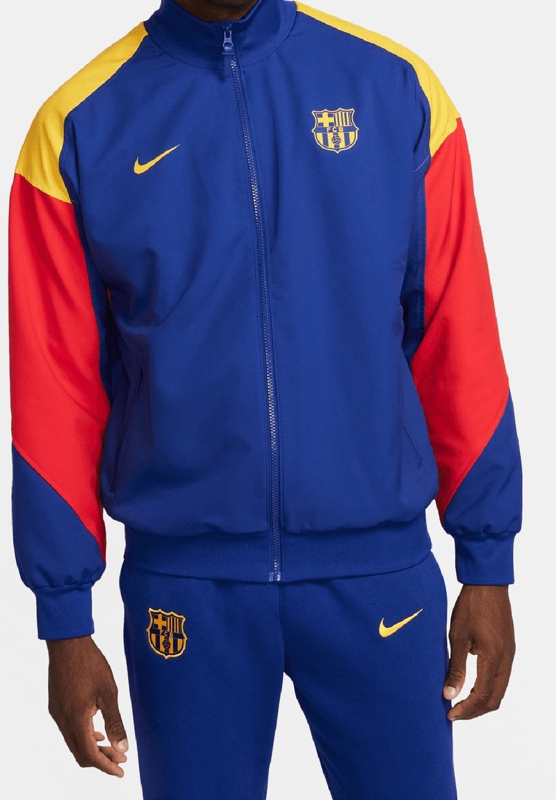 Chándal Nike azul y rojo con acentos amarillos. La chaqueta tiene un cierre de cremallera en la parte delantera, puños acanalados y el logo del FC Barcelona en el pecho.