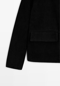 Schwarze Wildlederjacke mit einer rechteckigen Tasche auf der Vorderseite. Der Stoff wirkt weich und das Design hat lange Ärmel sowie einen geraden Saum.