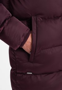 Giacca a vento bordeaux con una tasca con zip, polsini a costine, texture trapuntata e un sottile dettaglio del marchio vicino all'orlo.