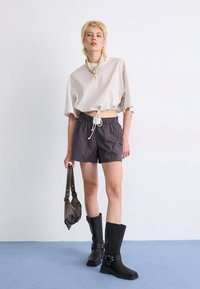 Lichtbeige gestreepte crop top met trekkoord, donkere gestreepte shorts, zwarte kniehoge laarzen en een kleine bruine tas. Soepele stoftexturen.