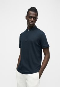 Jeune homme les mains dans les poches, portant un polo bleu marine à manches courtes côtelé et un pantalon blanc, regardant vers le bas et sur le côté.