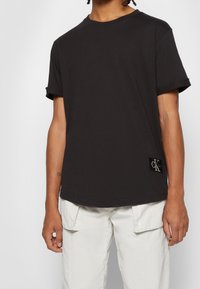 Man som bär en enkel svart t-shirt med kort ärm och en Calvin Klein-logotyp, samt vita cargobyxor med framfickor.