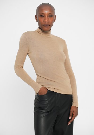 Pulover bej cu guler înalt, dintr-un material moale, cu un design ajustat, purtat cu pantaloni negri din piele, stând în fața unui fundal alb.