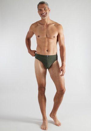 Lächelnder, oberkörperfreier Mann, der dunkelgrüne Badehosen mit weißen vertikalen Streifen trägt und barfuß vor einem schlichten hellen Hintergrund steht.