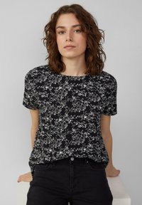 Zwart kortaf T-shirt met een wit bloemenpatroon, ronde halslijn en een losse pasvorm. G gedragen met zwarte jeans.