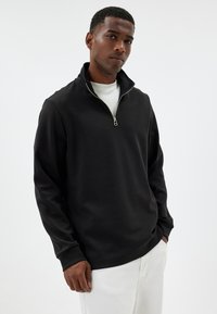 Black quarter-zip pullover made of soft fabric with a standing collar and long sleeves, paired with white trousers.