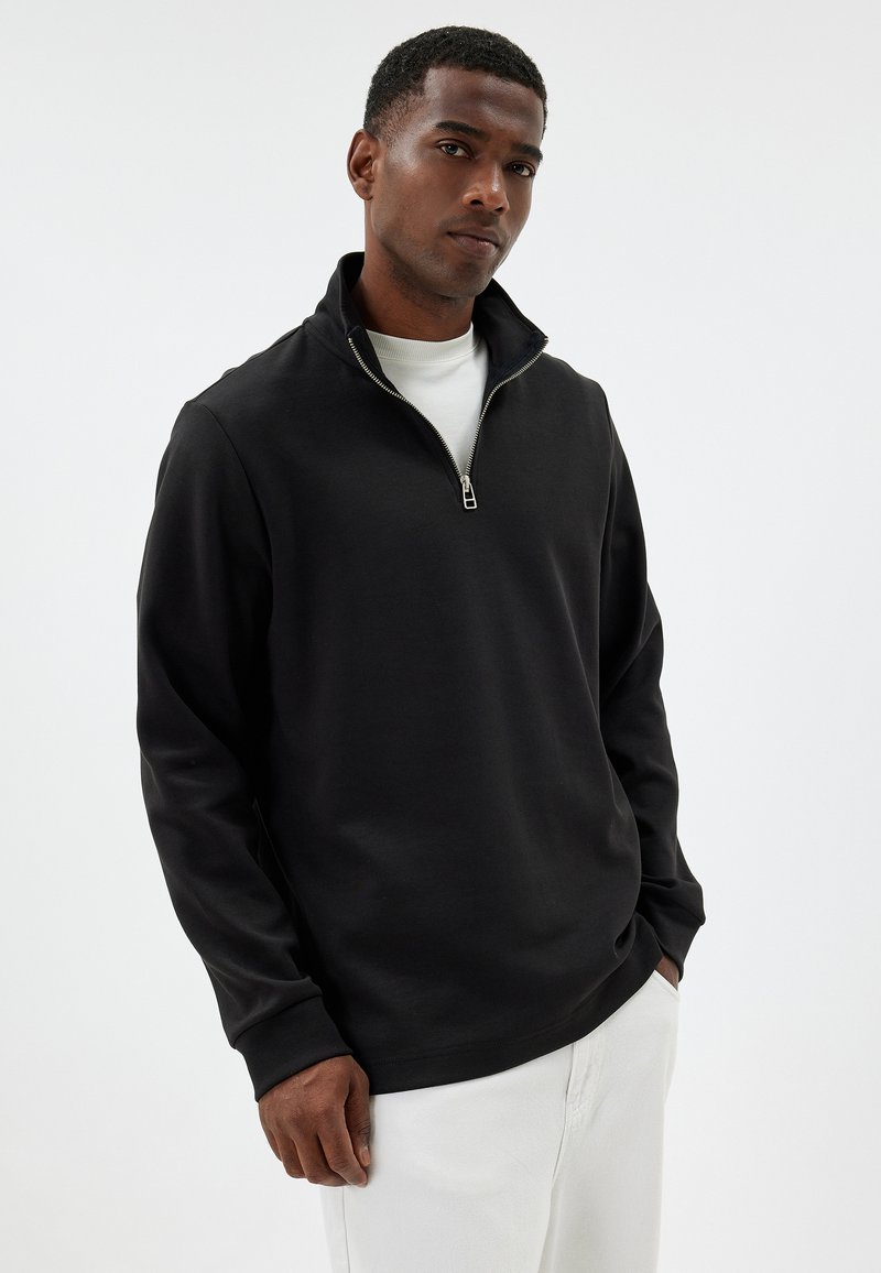 Black quarter-zip pullover made of soft fabric with a standing collar and long sleeves, paired with white trousers.