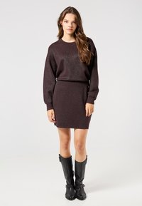 Dunkelbrauner gestrickter Pullover und passender Rock mit einem dezenten Schimmer. Das Model trägt kniehohe schwarze Lederstiefel. Schlichte, taillierte Silhouette.