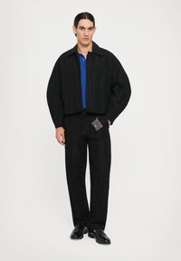 Schwarze, taillierte Jacke mit strukturiertem Muster, getragen über einem blauen Polohemd und passenden schwarzen Hosen. Enthält ein kleines angenähtes Etikett. Schwarze Schuhe.